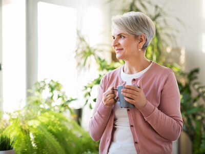 Frau steht entspannt mit einer Tasse in der Hand am Fenster und blickt ins Grüne – bewusste Pause im Pflegealltag | © PflegeBetreuer | unabhängige Pflegeberatung