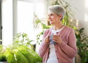 Frau steht entspannt mit einer Tasse in der Hand am Fenster und blickt ins Grüne – bewusste Pause im Pflegealltag | © PflegeBetreuer | unabhängige Pflegeberatung