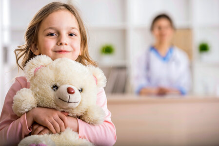 Kind mit Kuscheltier sitzt in einer Praxis während eine Fachkraft im Hintergrund sitzt | © PflegeBetreuer | unabhängige Pflegeberatung