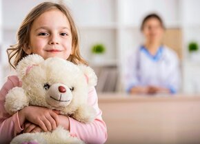 Kind mit Kuscheltier sitzt in einer Praxis während eine Fachkraft im Hintergrund sitzt | © PflegeBetreuer | unabhängige Pflegeberatung