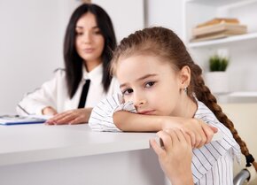 Mädchen lehnt mit dem Kopf auf einem Tisch, während im Hintergrund eine Fachperson sitzt | © PflegeBetreuer | unabhängige Pflegeberatung