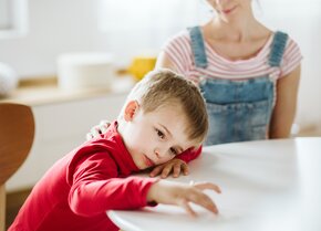 Kind sitzt nachdenklich am Tisch, während eine Frau unterstützend hinter ihm sitzt | © PflegeBetreuer | unabhängige Pflegeberatung