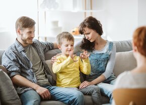 Kind sitzt mit seinen Eltern auf einem Sofa und zeigt einer Therapeutin eine Übung mit den Händen | © PflegeBetreuer | unabhängige Pflegeberatung