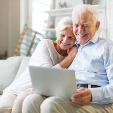 Älteres Paar liest auf dem Sofa sitzend gemeinsam einen Pflege-Newsletter am Laptop | © PflegeBetreuer | unabhängige Pflegeberatung