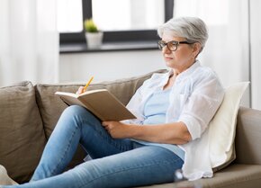 Ältere Frau sitzt entspannt auf dem Sofa und schreibt in ein Pflegetagebuch zur Dokumentation der Pflege | © PflegeBetreuer | unabhängige Pflegeberatung